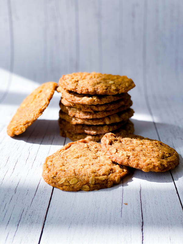 A stack of Anzac Biscuits with a couple in front on white boards.