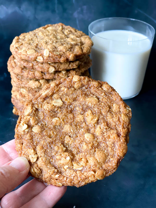 An Anzac Biscuit being held close up with more stacked in the background along with a glass of milk.