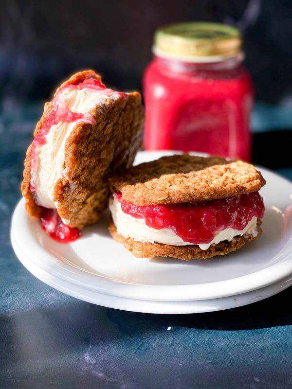 2 Anzac Biscuit ice-cream sandwiches with vanilla ice-cream and rhubarb compote sandwiched between 2 Anzac Biscuits with a jar of Rhubarb Compote in the background.