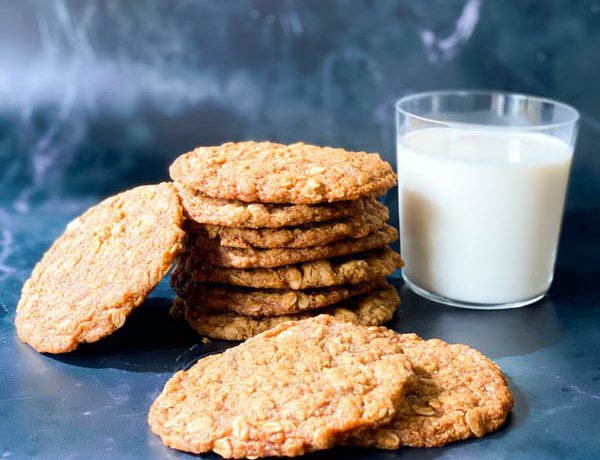 7 Anzac Biscuits stacked in a pile with 2 in front and one to the side with a glass of milk to the right on a lack marble surface.