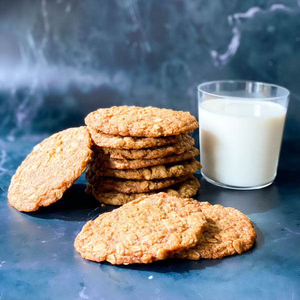 7 Anzac Biscuits stacked in a pile with 2 in front and one to the side with a glass of milk to the right on a lack marble surface.