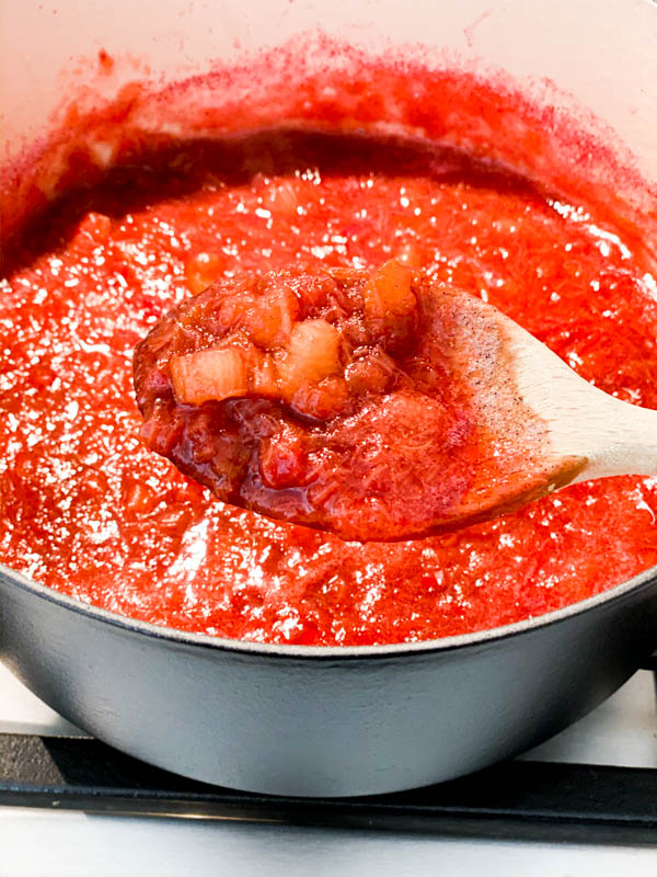 Rhubarb stewing in a pot with some lifted up in a wooden spoon showing the consistency with tiny pieces of rhubarb still present.