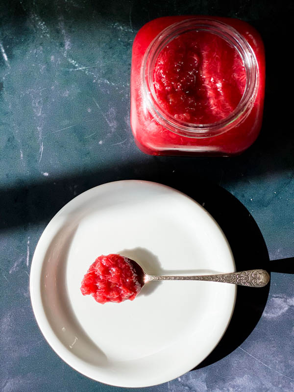 Looking down on a jar of Easy Rhubarb Compote and in front of it is a white side plate with a spoon of rhubarb resting on it.