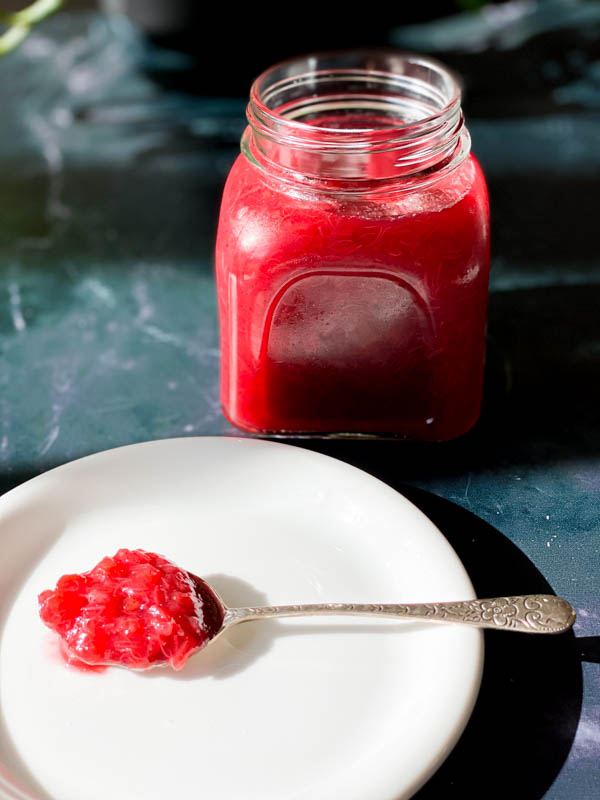 A side view of a jar of Easy Rhubarb Compote with a white plate in front that has a spoon filled with rhubarb compote.