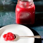 Close up of a jar of Easy Rhubarb Compote and in front of it is a white plate and spoon which has rhubarb compote on the spoon.