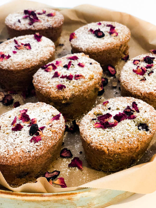 Pistachio Blueberry friands with icing sugar and rose petals on a platter lined with brown baking paper.