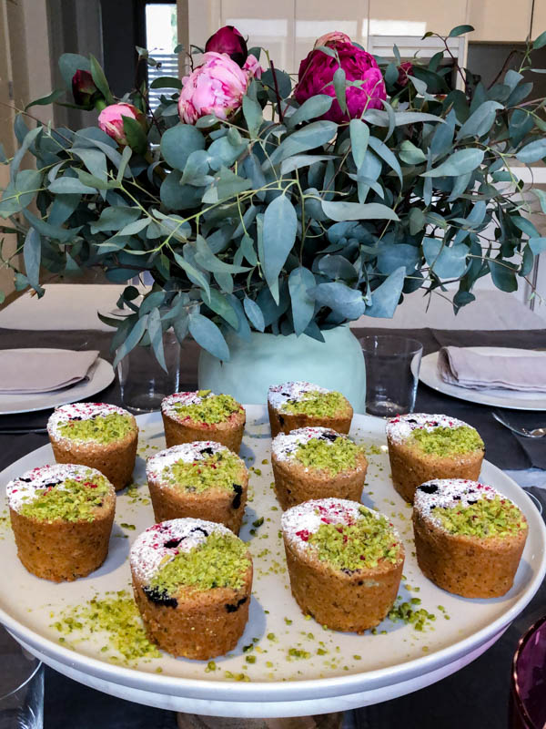 Friands on a white platter in front of a green vase with flowers on a dining table.