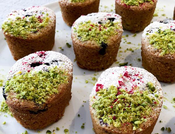 Pistachio Blueberry Friands on a white plate and decorated with Pistachio, Icing Sugar and curshed freeze-dried raspberries.