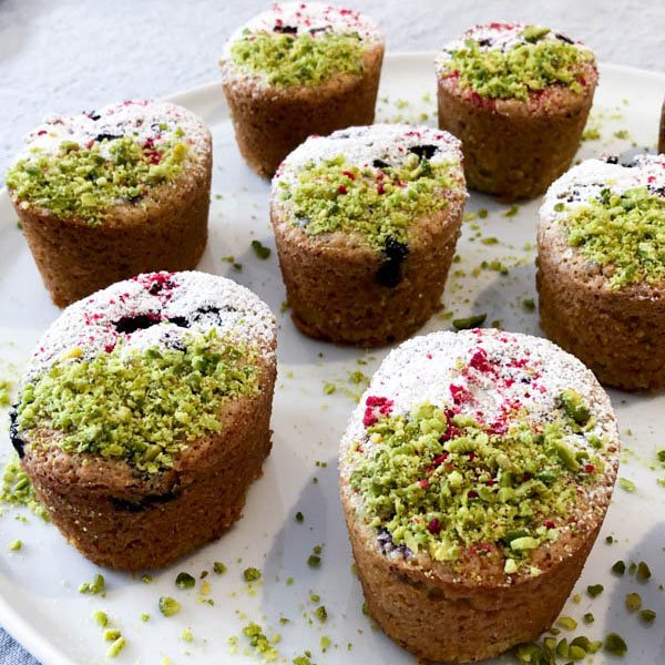 Pistachio Blueberry Friands on a white plate and decorated with Pistachio, Icing Sugar and curshed freeze-dried raspberries.