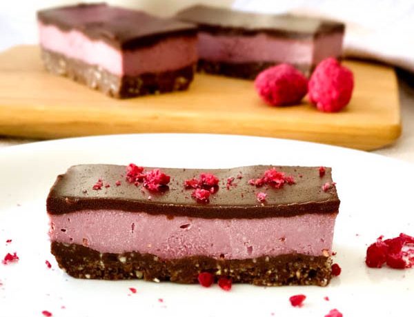 Close up of Raw Raspberry Chocolate Slice with freeze dried raspberries sprinkled around and 2 slices in background.