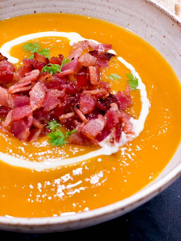 Close up of a bowl of Roasted Pumpkin Soup in a bowl topped with fried bacon pieces, parsley and a swirl of cream.