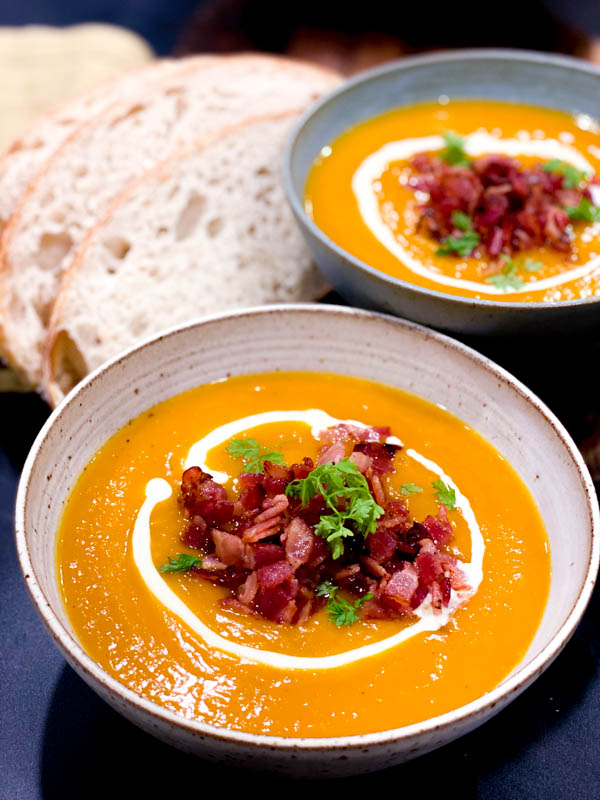 Roasted Pumpkin Soup in 2 bowls topped with fried bacon pieces, parsley and a swirl of cream with a side of fresh sourdough bread slices.