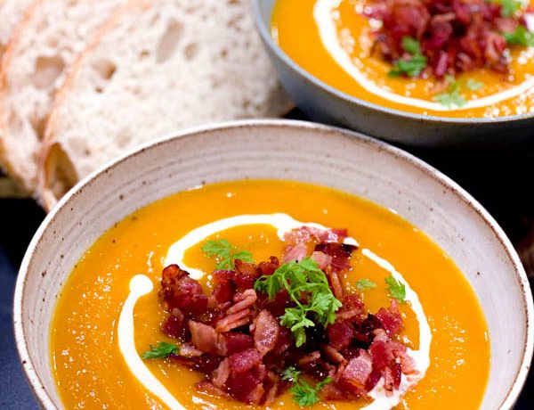 Close up of 2 bowls of Roasted Pumpkin Soup topped with bacon pieces, parsley and cream and served with a side of bread slices.