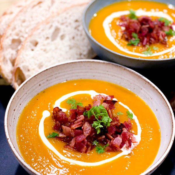 Close up of 2 bowls of Roasted Pumpkin Soup topped with bacon pieces, parsley and cream and served with a side of bread slices.