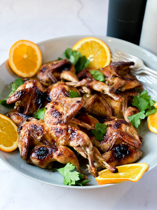 A platter of Butterflied Quail with an Orange, Cumin and Brown Sugar Glaze with orange slices around it.