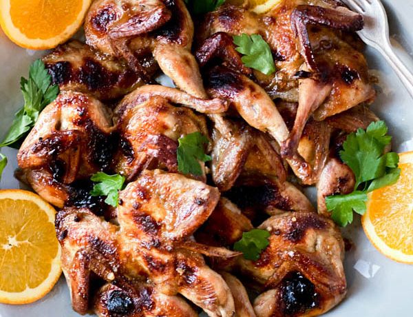 A platter of Butterflied Quail with an Orange, Cumin and Brown Sugar Glaze with orange slices around the outside and parsley leaves scattered over the quail. A couple of forks are on one side of the platter.