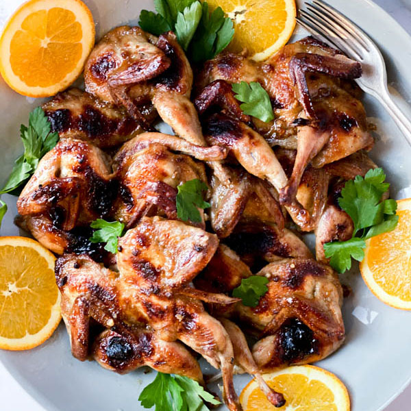 A platter of Butterflied Quail with an Orange, Cumin and Brown Sugar Glaze with orange slices around the outside and parsley leaves scattered over the quail. A couple of forks are on one side of the platter.