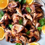 A round platter of Butterflied Quail with an Orange, Cumin and Brown Sugar Glaze. It has parsley leaves scattered over the top and a few orange slices around the quail. There are also 2 forks on one side of the dish.