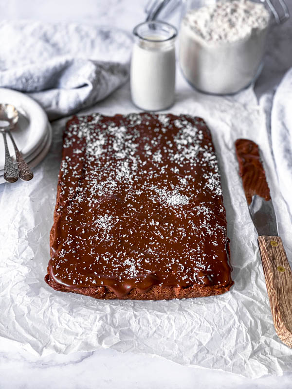 Looking down onto a slab of Chewy Chocolate Coconut Slice with a palette knife next to it with chocolate icing on it. Behind is a small jar of milk and a jar of flour with white dessert plates and spoons to the left.