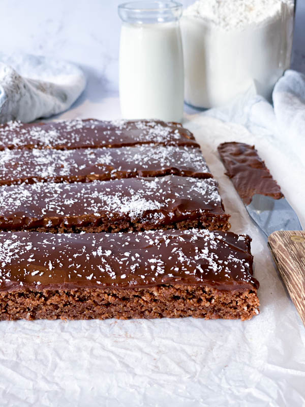 Close up of the sliced Chewy Chocolate Coconut Slice with a palette knife beside it and a small jar of milk behind it.