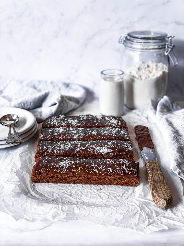 The Chewy Chocolate Coconut Slice on a sheet of crumpled white baking paper on a white marble benchtop. A palette knife with chocolate icing is beside it, with plates stacked to the left of it with 2 spoons on top. A large jar of flour and a small jar of milk are behind it.