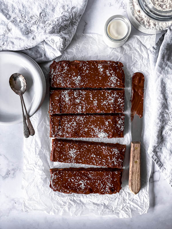 Looking down onto the Chewy Chocolate Coconut Slice with a palette knife to the right and plates and spoons to the left with a jar of milk behind it.