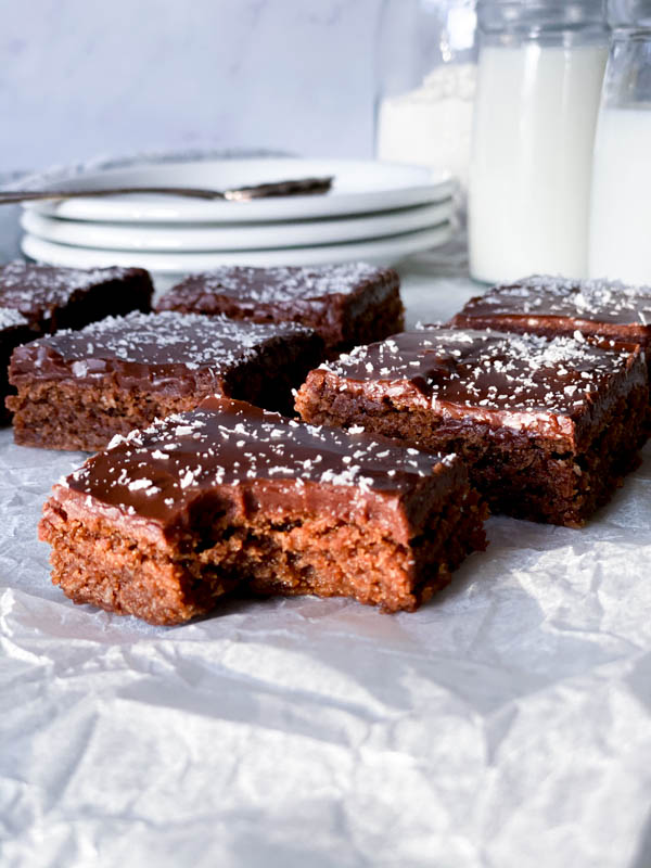 Close up of Chewy Chocolate Coconut Slices in squares with one bitten into at the front. In the background are a stack of 3 plates in the background.