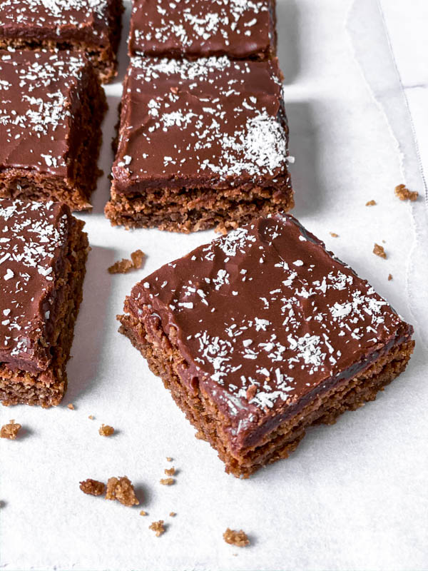 Chewy Chocolate Coconut Slices in squares with a few crumbs around it on white baking paper.