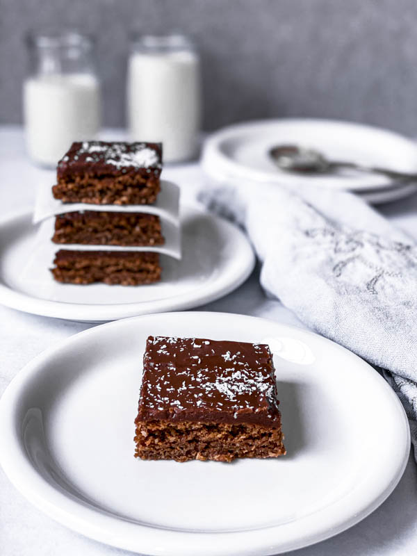 A Chewy Chocolate Coconut Slice piece on a white plate with 3 more pieces stacked on a plate behind it and a napkin beside it.