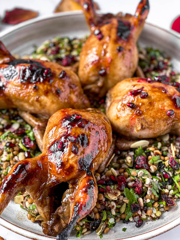 A round platter with the Cypriot Grain Salad underneath 4 whole Quail that are garnished with pomegranate seeds.