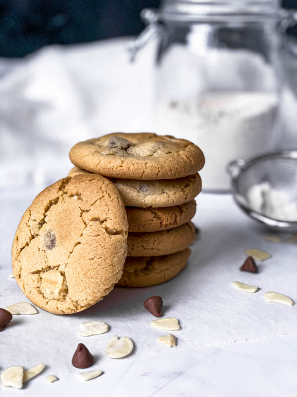 A stack of 5 Gingernut Biscuits with a 6th one leaning against them in the front. Around them scattered on the bench are almond flakes and a few chocolate nibs. In the background are a large jar of flour and a small sieve off to the right side partially out of view.