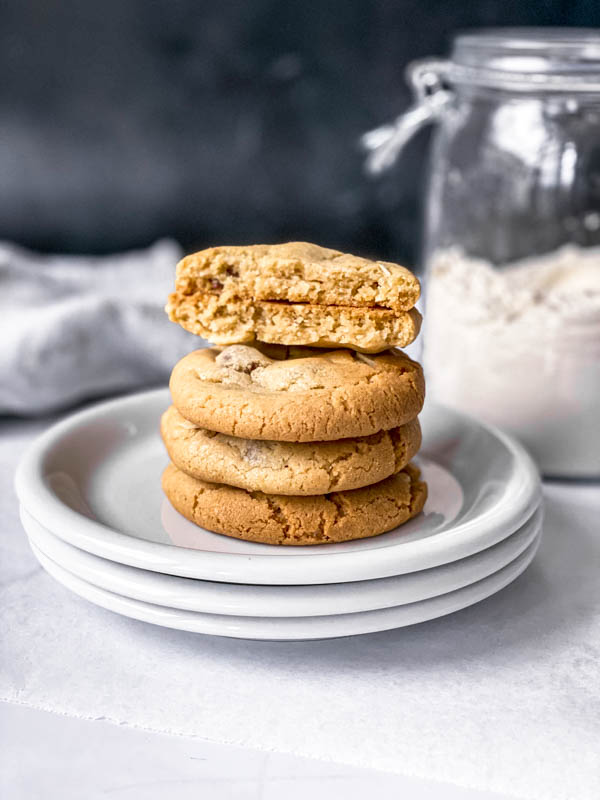 A stack of 3 whole Gingernut Biscuits with a fourth one broken in half on the top. They are stacked on 3 plates with a large jar of flour in the background.