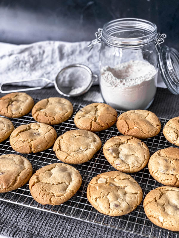 Gingernut Biscuits in 3 rows cooling on a wire cake resting rack with a jar of flour and a small sieve in the background. Everything is set on a dark grey tablecloth.