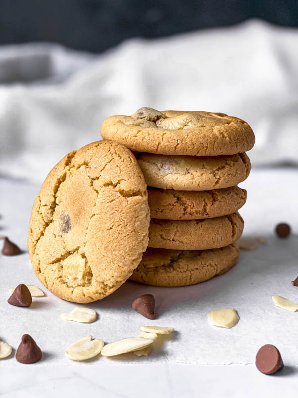 Close up of a stack of 5 Gingernut Biscuits with a 6th one leaning against them. Scattered around the front and sides of the stack are a few almond slices and milk chocolate nibs.