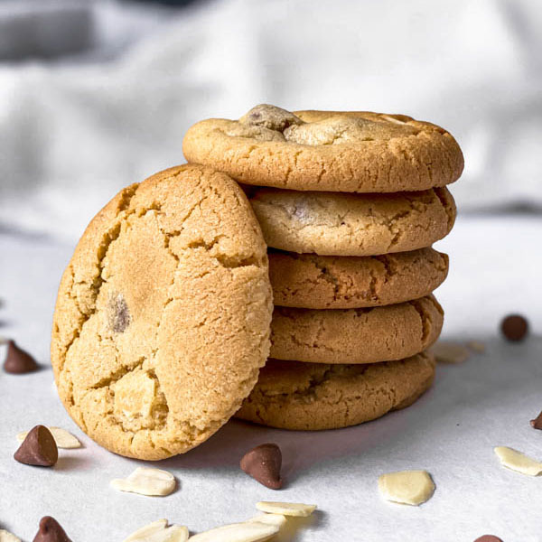 Close up of 5 stacked Gingernut Biscuits with one leaning against them. In the foreground are almond flakes and milk chocolate nibs.