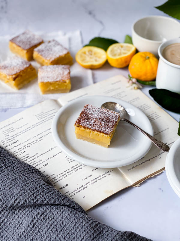 A square of Gooey Yuzu Slice on a white plate on top of an open cookbook with a spoon on the plate. There are also 4 more Gooey Yuzu squares behind as well as yuzu fruit and a cup of coffee.