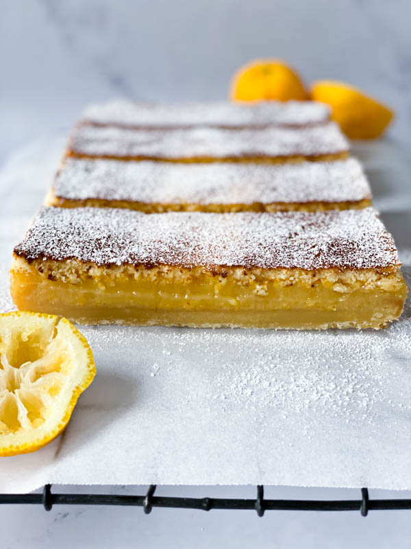 The Gooey Yuzu Slice is cut into long slices on white baking paper on top of a black resting rack with squeezed yuzu fruit in the front and back.