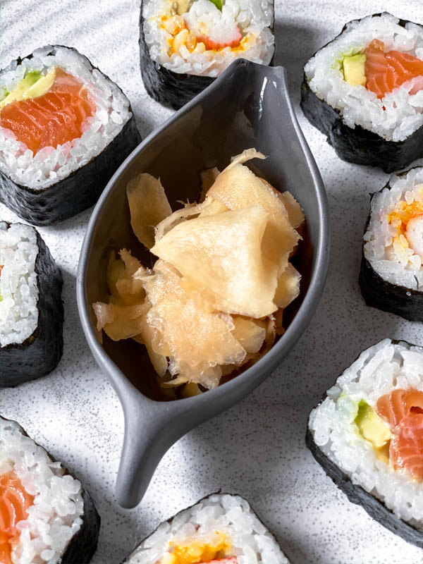 Some Easy Pickled Ginger in a small oval grey dish sitting in the middle of a large white platter surrounded by sushi rolls.