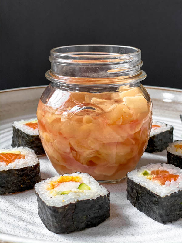 Looking front on at a jar of Easy Pickled Ginger with the lid off. It is in the middle of a large white pottery platter and has sushi rolls all around the jar.