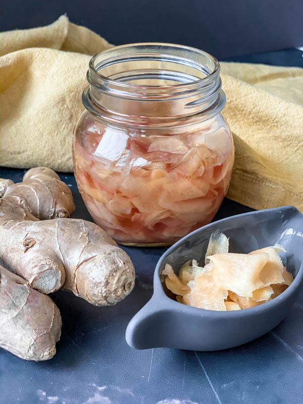 A jar of Easy Pickled Ginger with a knob of ginger in the foreground along with some Pickled Ginger in a small grey oval dish. In the background is a mustard coloured napkin.