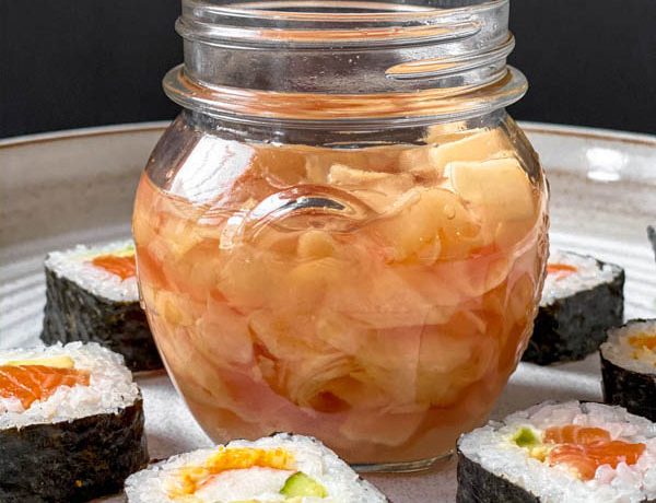 An open jar of Easy Pickled Ginger surrounded by cut sushi rolls on a large white ceramic platter.