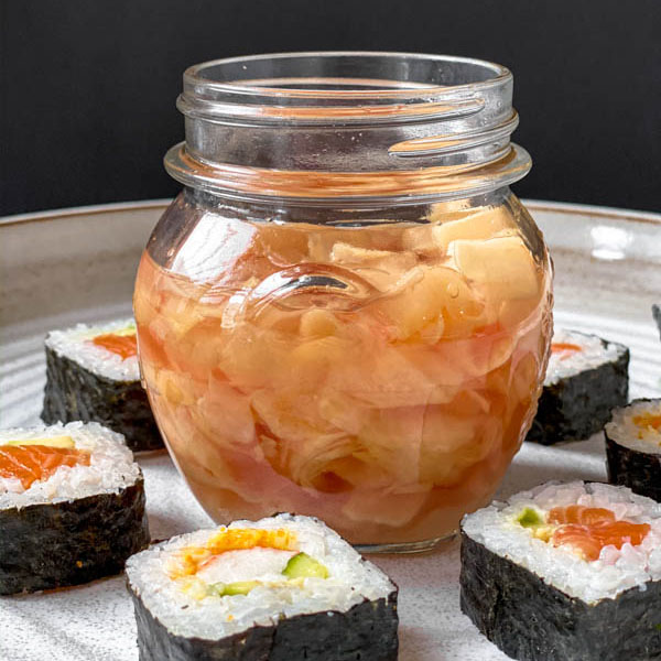 An open jar of Easy Pickled Ginger surrounded by cut sushi rolls on a large white ceramic platter.