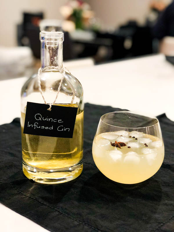 A bottle of Quince Infused Gin with a glass of poured gin with ice, star anise, black peppercorns and tonic sitting beside it.