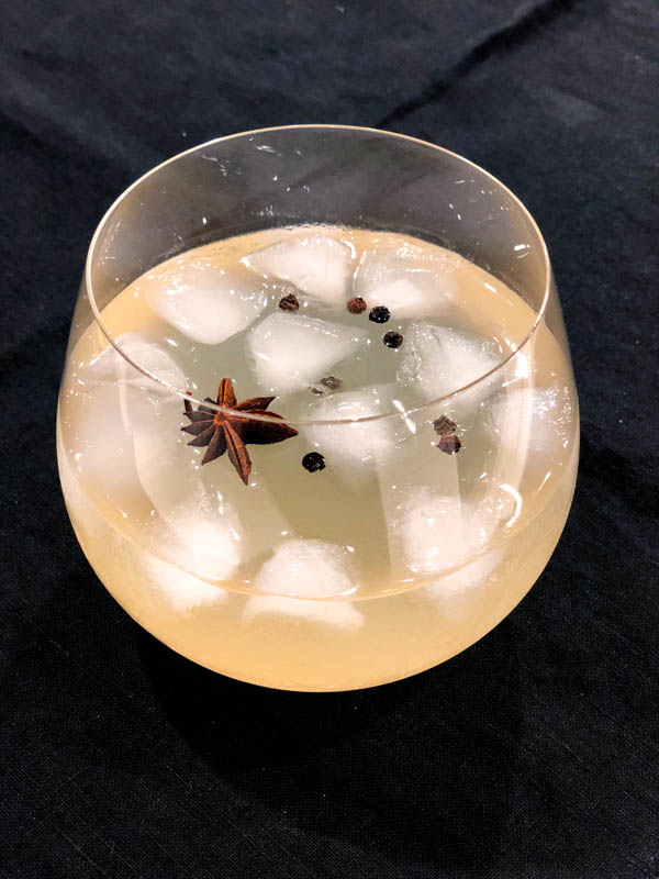 Side view of the glass of Quince Infused Gin with tonic water, ice, star anise and black peppercorns on a dark tablecloth.