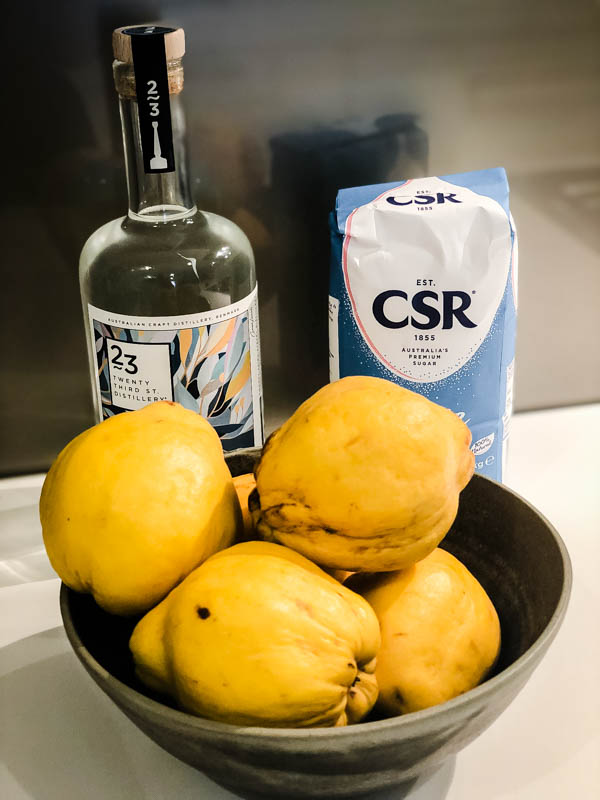 A bottle of Gin, packet of sugar, and bowl of washed Quince fruit on a benchtop.
