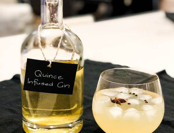 A bottle of Quince Infused Gin with a glass of poured gin and tonic beside it on a white bench.