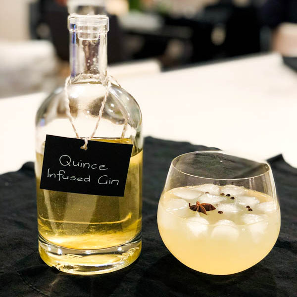 A bottle of Quince Infused Gin with a glass of poured gin and tonic beside it on a white bench.