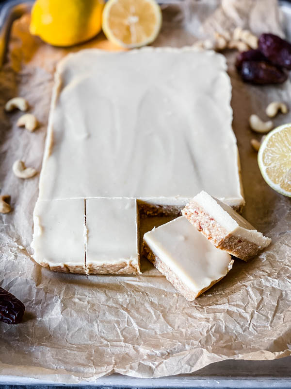 The Raw Lemon Slice shown mostly unsliced, on brown baking paper with dates, cashews and lemons surrounding the slice.