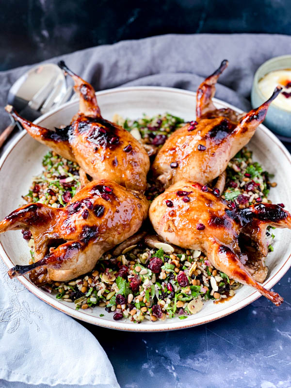 The white round ceramic platter with Roasted Pomegranate Quail on top of a salad with grey napkins around it as well as salad servers and a small bowl of yoghurt dressing to the sides.