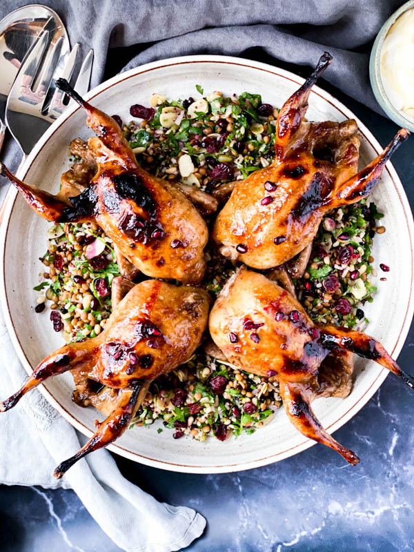 Looking down onto the platter of Roasted Pomegranate Quail on a dark marble table with grey napkins, salad servers around it.